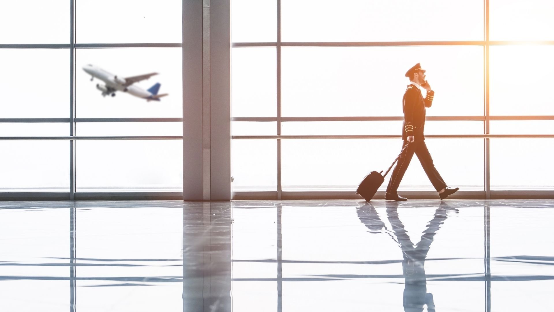Pilot in uniform pulling luggage watches airplane take off through airport window.